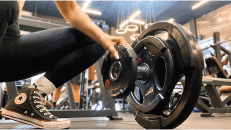 Mão ajustando anilhas em uma barra de musculação na academia, com tênis All Star no chão.